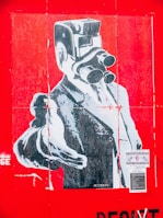 A stylized poster features a surreal image of a person in formal attire with an old-fashioned camera in place of their head. The entire image is dominated by a bright red background. The person is wearing a suit and pointing forward, giving a strong and commanding presence. There are several pieces of text and stickers on the poster, adding to the urban and artistic feel.