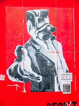 A stylized poster features a surreal image of a person in formal attire with an old-fashioned camera in place of their head. The entire image is dominated by a bright red background. The person is wearing a suit and pointing forward, giving a strong and commanding presence. There are several pieces of text and stickers on the poster, adding to the urban and artistic feel.