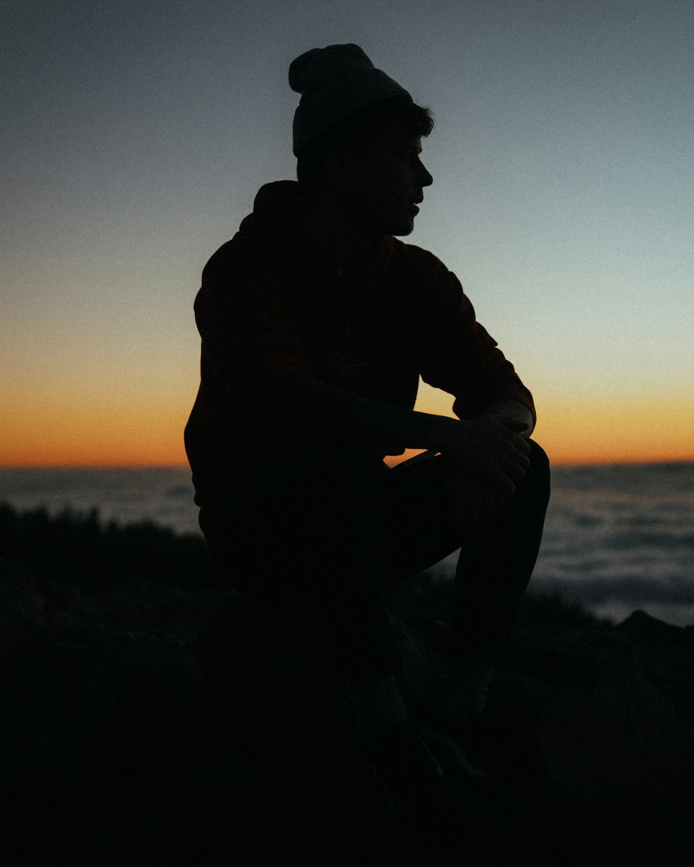 silhouette of man wearing jacket and cap sitting on rock during sunset