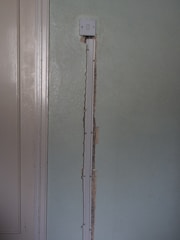 A partially exposed light switch is mounted on a light green textured wall, with visible wiring and a vertical conduit extending downward. The wall has noticeable indentations and unfinished edges where the conduit is installed.