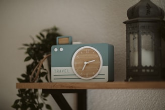 A decorative clock designed to resemble a vintage camera sits on a wooden shelf. The clock features a circular face with wooden texture and white hands, and the word 'TRAVEL' written on its front. Nearby, a dark, ornate lantern stands, and a leafy green plant adds a natural touch in the background.