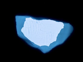 A large, irregularly shaped iceberg is surrounded by dark water, creating a striking contrast. The iceberg features intricate patterns and textures across its surface, suggesting a weathered and natural form.