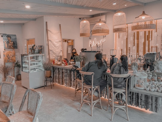 A cozy caf&eacute; interior with a rustic and bohemian decor style, featuring rattan chairs and pendant lights, a counter with a barista, and patrons sitting on high stools. The walls are adorned with artworks and potted plants, creating a warm and inviting atmosphere.