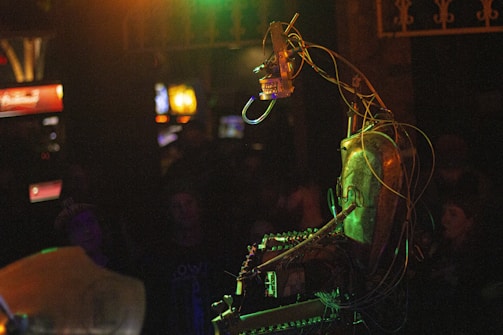 A mechanical structure with an arrangement of wires and metal components is illuminated by colorful lights. The setting appears to be a dimly lit room, possibly a bar or club, with blurred figures in the background.