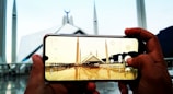 A pair of hands holding a smartphone that is taking a picture of a large mosque with multiple minarets. The phone screen shows a clearer view of the mosque, with the sky and mountains in the background.