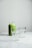 A glass jar filled with green smoothie is placed on a white marble surface, accompanied by two empty clear glasses. The background is softly lit, giving the scene a clean and minimalistic appearance.