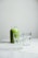 A glass jar filled with green smoothie is placed on a white marble surface, accompanied by two empty clear glasses. The background is softly lit, giving the scene a clean and minimalistic appearance.