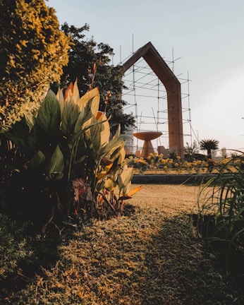 A beautifully landscaped garden with lush green foliage in the foreground, orange and yellow flowers, and a large architectural structure with scaffolding in the background. The sunlight casts a warm glow over the entire scene, creating contrasting shadows on the grass.