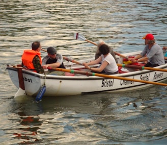 A group of people rowing a boat labeled 'Bristol Channel Social Rowers' on a body of water. One person wears an orange life jacket, while others are dressed casually. They hold oars, coordinating their movements.