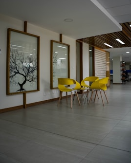A modern interior space with three bright yellow chairs and small round tables on a sleek tiled floor. The walls are decorated with framed glass or acrylic panels featuring tree silhouettes. The ceiling showcases a combination of recessed lighting and wooden slats, contributing to a contemporary design. The area appears to be minimalistic and open, likely part of an office or lounge.