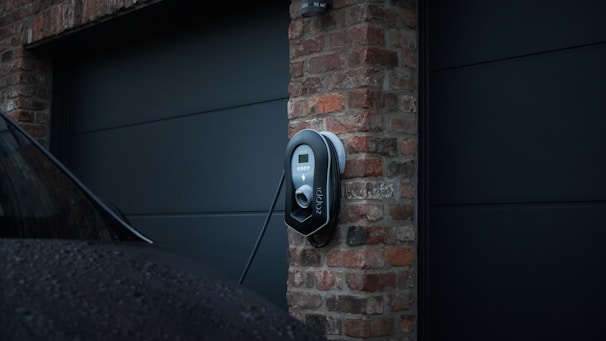 A modern electric vehicle charging station is mounted on a brick wall in a residential setting. The charger appears sleek and is connected to a vehicle parked nearby. The lighting suggests an overcast day, with the brick wall providing a rustic contrast to the contemporary design of the charger.
