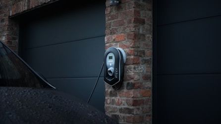 A modern electric vehicle charging station is mounted on a brick wall in a residential setting. The charger appears sleek and is connected to a vehicle parked nearby. The lighting suggests an overcast day, with the brick wall providing a rustic contrast to the contemporary design of the charger.