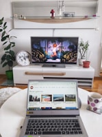 A modern living room setup with a flat-screen TV displaying an image of a woman in a white outfit surrounded by decorative elements. In the foreground, a laptop is open on a white round table, showing a webpage from CNET featuring photo galleries. A polka-dotted mug is next to the laptop. Various houseplants are arranged in the background, along with a large wall clock and decorative items on a shelf.
