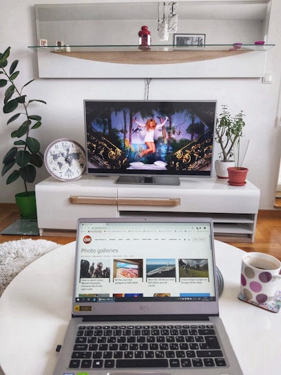 A modern living room setup with a flat-screen TV displaying an image of a woman in a white outfit surrounded by decorative elements. In the foreground, a laptop is open on a white round table, showing a webpage from CNET featuring photo galleries. A polka-dotted mug is next to the laptop. Various houseplants are arranged in the background, along with a large wall clock and decorative items on a shelf.
