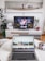 A modern living room setup with a flat-screen TV displaying an image of a woman in a white outfit surrounded by decorative elements. In the foreground, a laptop is open on a white round table, showing a webpage from CNET featuring photo galleries. A polka-dotted mug is next to the laptop. Various houseplants are arranged in the background, along with a large wall clock and decorative items on a shelf.
