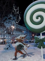A whimsical winter scene features a large mouse statue adorned with a scarf and skates, set among oversized lollipop decorations in a snowy outdoor setting. Festive lights illuminate nearby trees and buildings, creating a cheerful and lively atmosphere.
