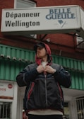 A person stands outside a building wearing a black jacket with red accents and a red hood. They are looking to the side, adjusting the front of the jacket with their hands. The building has a sign that reads 'Dépanneur Wellington' and 'Belle Gueule'. The structure is made of red brick with a green and white awning.