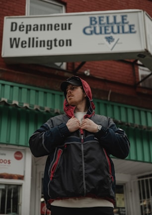 A person stands outside a building wearing a black jacket with red accents and a red hood. They are looking to the side, adjusting the front of the jacket with their hands. The building has a sign that reads 'Dépanneur Wellington' and 'Belle Gueule'. The structure is made of red brick with a green and white awning.
