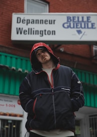 A person wearing a dark hooded jacket with red details stands in front of a sign for 'Dépanneur Wellington' and 'Belle Gueule'. The background features a building with a green awning and a partially visible storefront window.