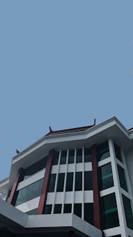 A modern multi-story building with a unique architectural design featuring large windows and bold vertical and horizontal lines. The structure is composed of a combination of white and red colors, with a roof that has distinct overhangs and possibly Asian influences. The sky in the background is clear and blue, creating a peaceful and open atmosphere.