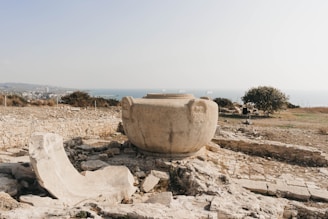 An ancient stone vessel, possibly used in past civilizations, is situated among ruins on a hilly area. The background features a distant coastline and a cityscape partially obscured by the hazy atmosphere. Sparse vegetation and scattered stones add to the arid, historical setting.