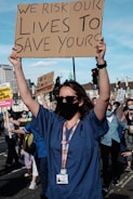 A person wearing a blue medical uniform and a black mask holds up a cardboard sign that reads, 'We risk our lives to save yours.' The background features other people also wearing masks and holding signs, with buildings and traffic lights visible.