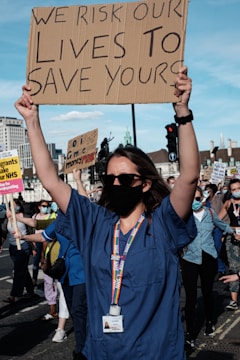 A person wearing a blue medical uniform and a black mask holds up a cardboard sign that reads, 'We risk our lives to save yours.' The background features other people also wearing masks and holding signs, with buildings and traffic lights visible.