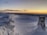 A snow-covered landscape with a fence partially buried in snow stretches into the distance. A survey or trig point made of stone stands prominently in the foreground. The sky is clear with a palette of warm yellows and oranges near the horizon, transitioning to a darker blue as it rises, indicating either sunrise or sunset.