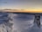 A snow-covered landscape with a fence partially buried in snow stretches into the distance. A survey or trig point made of stone stands prominently in the foreground. The sky is clear with a palette of warm yellows and oranges near the horizon, transitioning to a darker blue as it rises, indicating either sunrise or sunset.