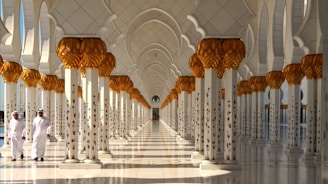 An ornate corridor with an intricately designed ceiling and columns adorned with golden palm motifs. Two individuals in traditional Middle Eastern attire walk along the polished marble floor, casting subtle reflections.