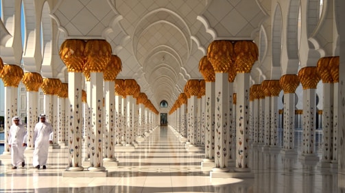 An ornate corridor with an intricately designed ceiling and columns adorned with golden palm motifs. Two individuals in traditional Middle Eastern attire walk along the polished marble floor, casting subtle reflections.