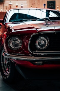 A close-up of a classic red car with prominent round headlights and chrome accents, parked indoors with trophies displayed in the background on wooden walls.