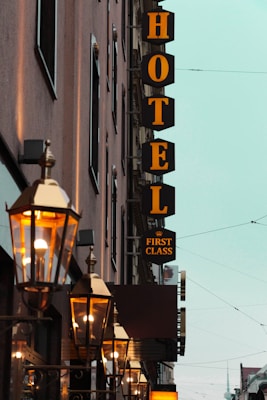 A row of illuminated vintage-style street lamps lines the side of a building with a prominent sign displaying 'HOTEL' and 'FIRST CLASS' in bold yellow letters against a dark background. The facade of the building is captured at an angle, giving a sense of depth with additional architectural features and windows visible. The sky is a pale blue-green shade, contributing to an evening or late afternoon atmosphere.