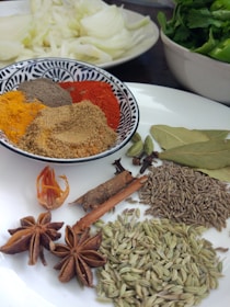 Various spices are arranged on a white plate and in a decorative bowl. The bowl contains colorful ground spices, including yellow turmeric, brown cumin, and red chili powder. On the plate are whole spices such as star anise, cinnamon sticks, fennel seeds, bay leaves, and cardamom pods. In the background, there is a plate with sliced onions and a bowl of green leafy vegetables.
