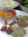 Various spices are arranged on a white plate and in a decorative bowl. The bowl contains colorful ground spices, including yellow turmeric, brown cumin, and red chili powder. On the plate are whole spices such as star anise, cinnamon sticks, fennel seeds, bay leaves, and cardamom pods. In the background, there is a plate with sliced onions and a bowl of green leafy vegetables.