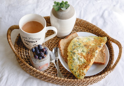 A wicker tray holds a breakfast arrangement including a cup of coffee with a message, a ceramic plant pot, a plate with an omelette on toast, and a jar of chia pudding topped with blueberries. A spoon is also present.