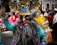 A person wearing an elaborate and colorful costume with intricate details. The vibrant outfit includes a headdress with feathers and decorative elements, and the face is adorned with a mask featuring exaggerated features. In the background, other people also dressed in traditional costumes are visible, suggesting a parade or cultural event.