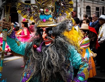 A person wearing an elaborate and colorful costume with intricate details. The vibrant outfit includes a headdress with feathers and decorative elements, and the face is adorned with a mask featuring exaggerated features. In the background, other people also dressed in traditional costumes are visible, suggesting a parade or cultural event.