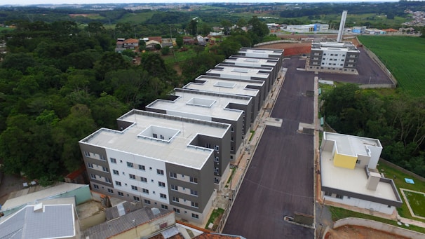 Aerial view of a residential complex featuring multiple uniform apartment buildings with flat roofs. The buildings are arranged in a row alongside a newly paved road. Surrounding the complex are areas of lush greenery and fields, with a few scattered houses visible in the distance. The overall landscape combines urban development with natural elements.