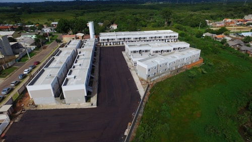 Aerial view of a residential area with a series of connected white buildings forming a housing complex. The complex is surrounded by a mix of paved roads and open green spaces with a few scattered houses nearby. A water tower is visible in the background, alongside other residential buildings.