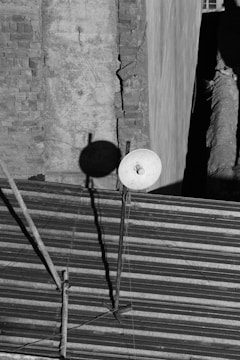 A black and white image depicts a rooftop with a corrugated metal surface. There is a satellite dish mounted on a pole, casting a shadow on a weathered brick wall backdrop. The wall shows signs of wear with visible cracks.