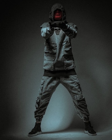 A figure wearing a hooded jacket and cargo pants stands in a dimly lit environment. The face is partially obscured by the hood, and the gloves cover the hands in a pointing gesture. The clothing is adorned with pockets and a prominent skull design on the chest.