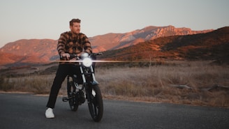 A person rides a modern electric motorcycle on a paved road set against a backdrop of rugged mountains bathed in warm, golden light. The grass on the roadside is dry, and the overall scenery suggests a serene, rural environment during sunset or sunrise.