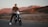A person rides a modern electric motorcycle on a paved road set against a backdrop of rugged mountains bathed in warm, golden light. The grass on the roadside is dry, and the overall scenery suggests a serene, rural environment during sunset or sunrise.