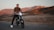 A person rides a modern electric motorcycle on a paved road set against a backdrop of rugged mountains bathed in warm, golden light. The grass on the roadside is dry, and the overall scenery suggests a serene, rural environment during sunset or sunrise.