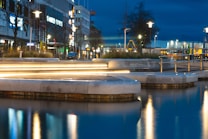 A nighttime urban scene features a well-lit street with modern buildings and streetlamps reflecting off a body of water in the foreground. Light trails from passing vehicles create a dynamic, glowing line across the image. Sparse trees and various signs add to the city atmosphere.