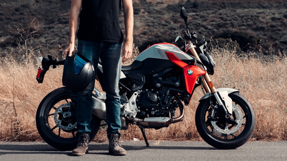 man in black t-shirt and blue denim jeans standing beside red and black sports bike