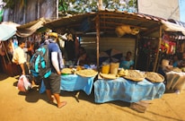 A bustling outdoor market scene featuring a vendor stall with various grains and pulses displayed in large baskets on a blue tablecloth. The vendor, dressed in a blue sari, sits behind the stall with an umbrella providing shade. Several people, including a man with a backpack and hat, are engaged in activity nearby. Bright colors and textiles can be seen hanging in the background.