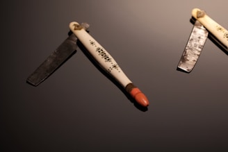 Two vintage straight razors are placed on a dark reflective surface. The handles are ornately decorated and feature intricate patterns. One razor is open, revealing a polished blade, while the other is only partially visible. The lighting highlights the textures of both the metal and the handle.