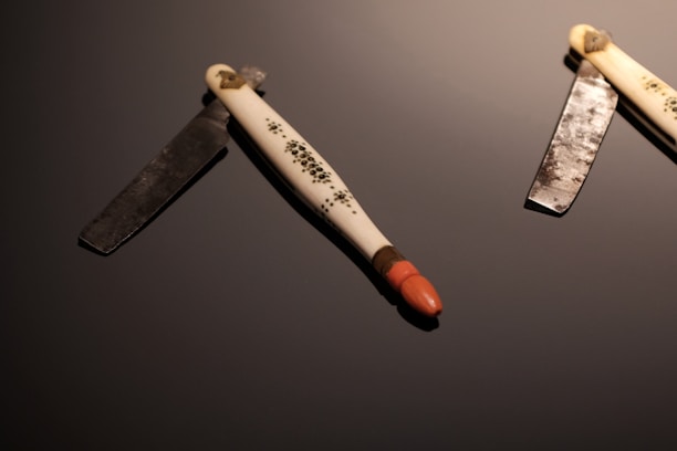 Two vintage straight razors are placed on a dark reflective surface. The handles are ornately decorated and feature intricate patterns. One razor is open, revealing a polished blade, while the other is only partially visible. The lighting highlights the textures of both the metal and the handle.
